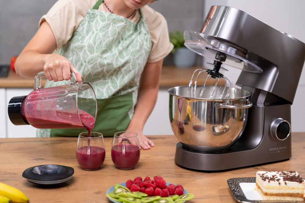 Osoba w zielonym fartuchu nalewa czerwony napój z blendera do dwóch szklanek na blacie kuchennym. Obok znajduje się robot kuchenny planetarny z otwartą pokrywą i widocznymi trzepaczkami, a na blacie leżą owoce i ciasto.