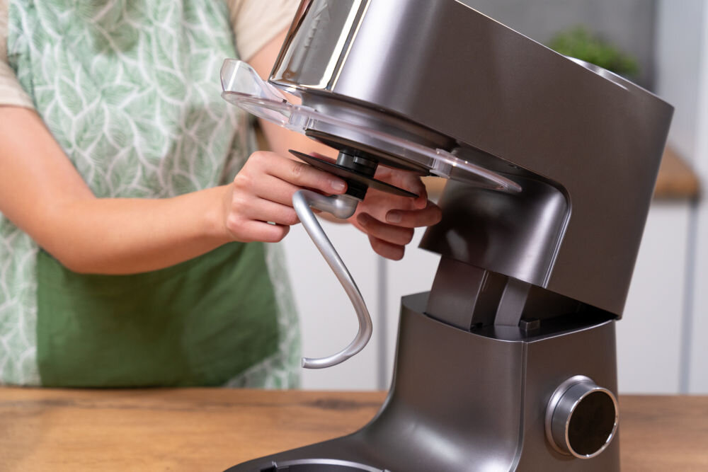 Osoba montuje hak do mieszania w robocie kuchennym planetarnym o metalicznym wykończeniu. W tle widoczny jest fragment kuchni.