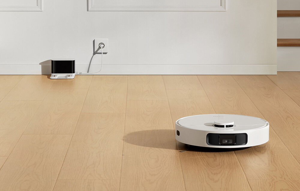Na drewnianej podłodze znajduje się biały robot sprzątający o okrągłym kształcie. W tle widać stację dokującą podłączoną do gniazdka elektrycznego.