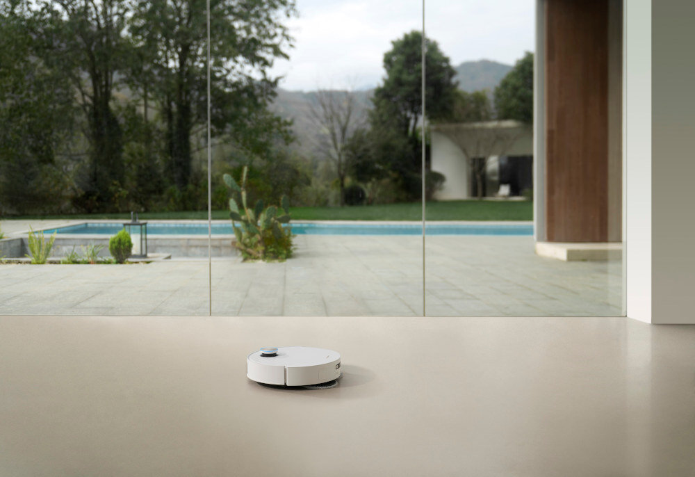 Na podłodze w pomieszczeniu znajduje się biały robot sprzątający. W tle widoczny jest duży przeszklony taras z widokiem na basen i ogród.