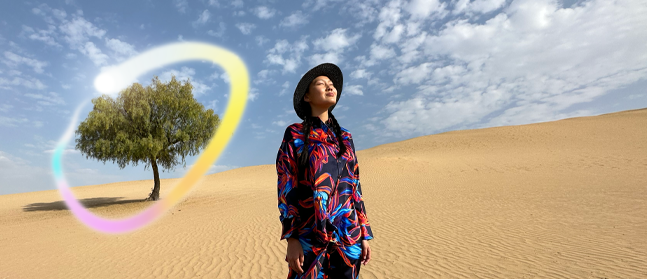 Osoba w kolorowym stroju i kapeluszu stoi na pustyni, patrząc w górę. W tle widoczna jest samotna zielona roślina oraz kolorowy pierścień świetlny.
