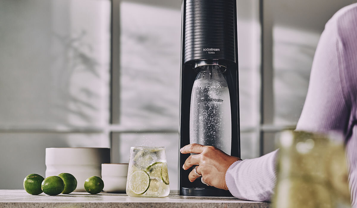Osoba używa czarnego saturatora do gazowania wody, na którym widnieje napis \'sodastream Terra\'. Obok na blacie znajdują się limonki i szklanka z napojem oraz plasterkami limonki.