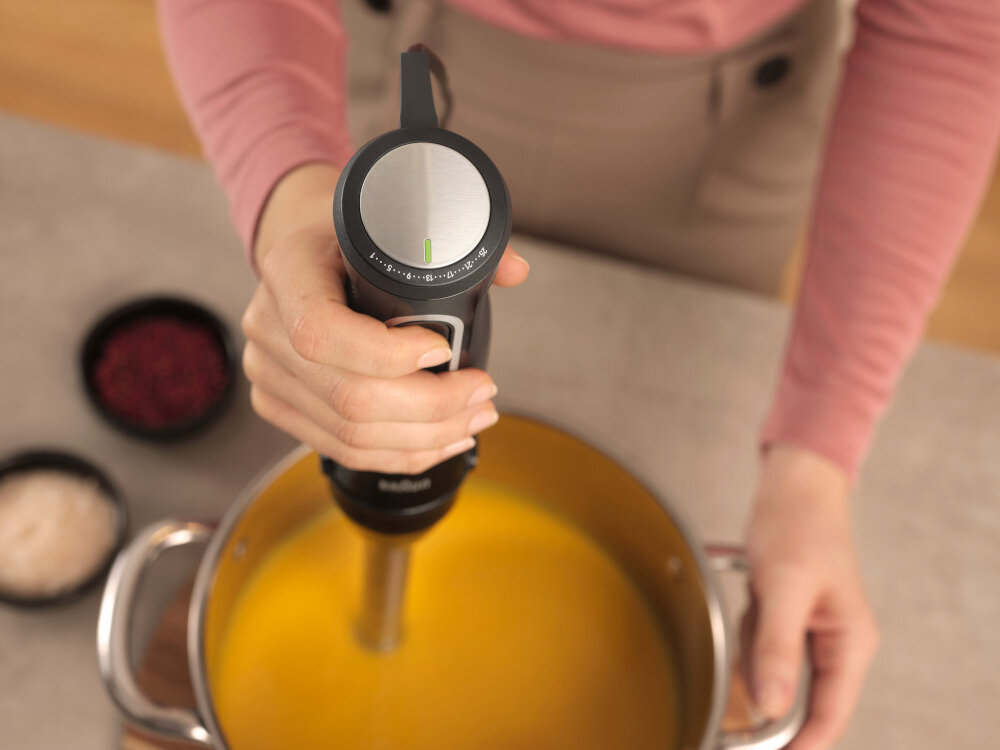 Osoba trzyma blender zanurzeniowy nad garnkiem z żółtą zupą. Na blacie widać dwie miseczki z przyprawami, a na blenderze widoczne jest logo \'Braun\'.