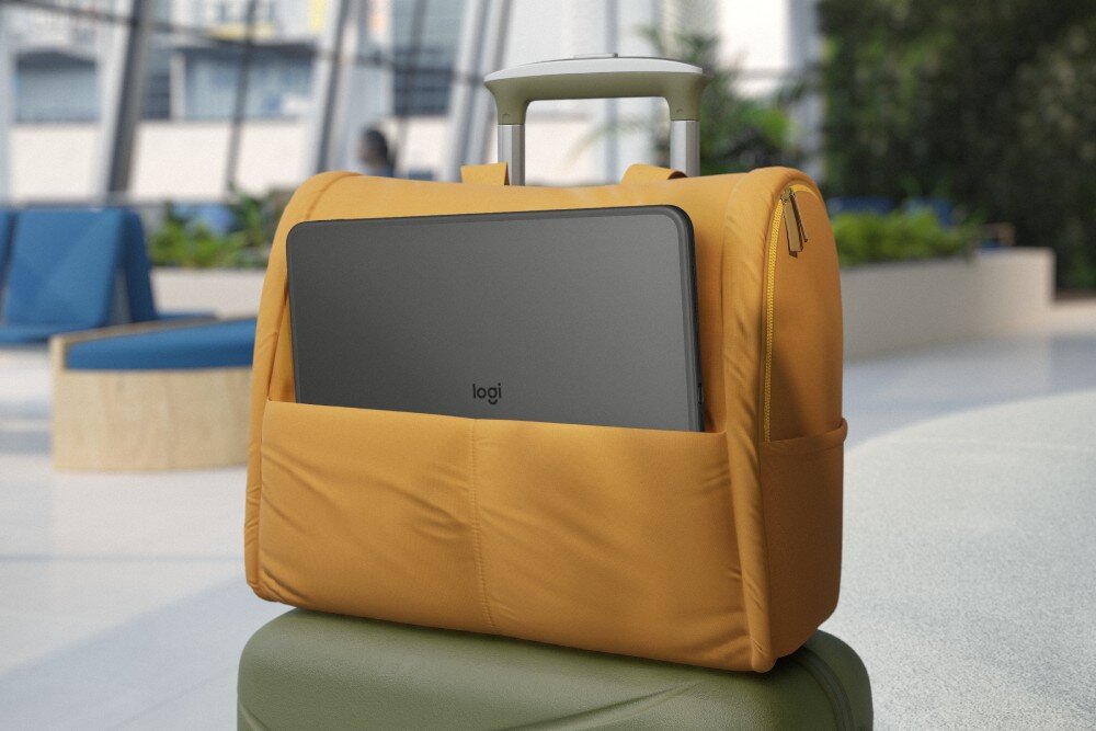 Na obrazie widać żółtą torbę podróżną umieszczoną na walizce, z wystającym z niej urządzeniem z logo \'logi\'. W tle znajduje się wnętrze budynku z roślinami i siedzeniami.