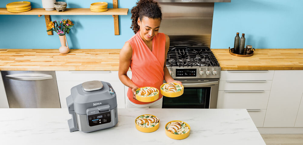 Kobieta w kuchni trzyma talerz z jedzeniem, obok niej na blacie stoi frytkownica beztłuszczowa z widocznym logo \'NINJA\'.