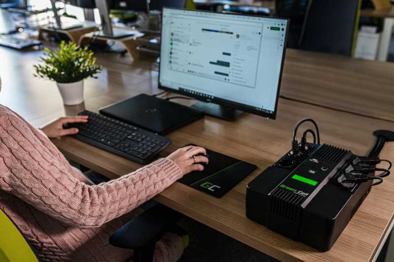 Osoba siedzi przy biurku z komputerem, obok którego znajduje się zasilacz UPS z widocznym logo Green Cell. Na ekranie monitora wyświetlona jest aplikacja komunikacyjna.