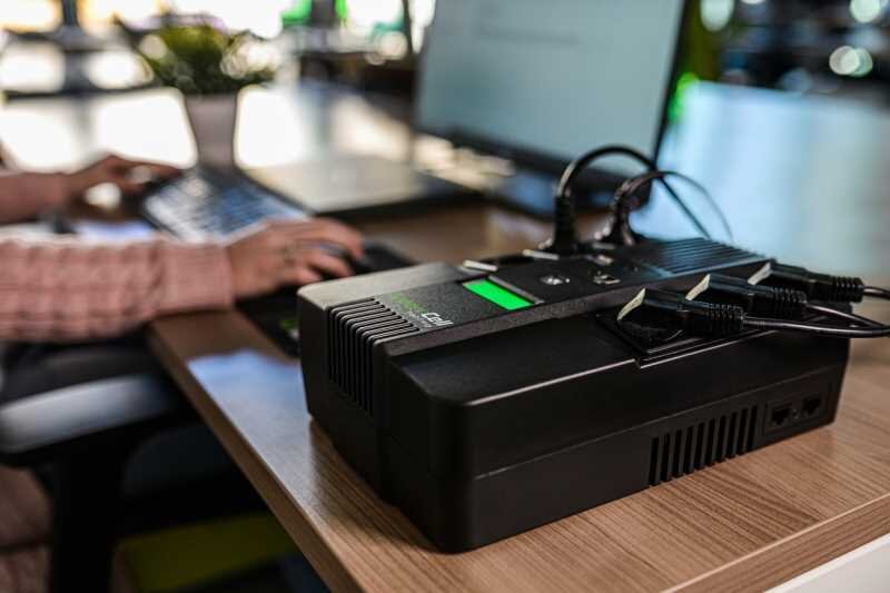 Na biurku znajduje się czarny zasilacz UPS z kilkoma podłączonymi kablami. W tle widoczna jest osoba pracująca przy laptopie.