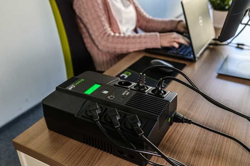 Na biurku znajduje się zasilacz UPS z logo Green Cell, do którego podłączone są różne przewody. W tle osoba pracuje na laptopie.