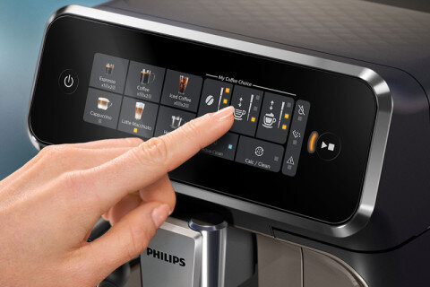Osoba dotyka ekranu dotykowego ekspresu do kawy, na którym widoczne są różne opcje napojów, w tym \'Iced Coffee\'. Na urządzeniu znajduje się logo \'PHILIPS\'.
