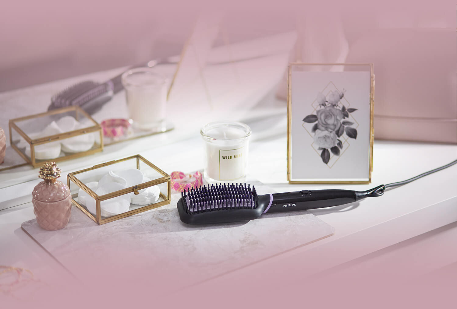 Na białej powierzchni leży szczotka prostująca z napisem \'PHILIPS\', obok której znajduje się świeca z napisem \'WILE\'. W tle widoczna jest ramka z grafiką przedstawiającą kwiaty.