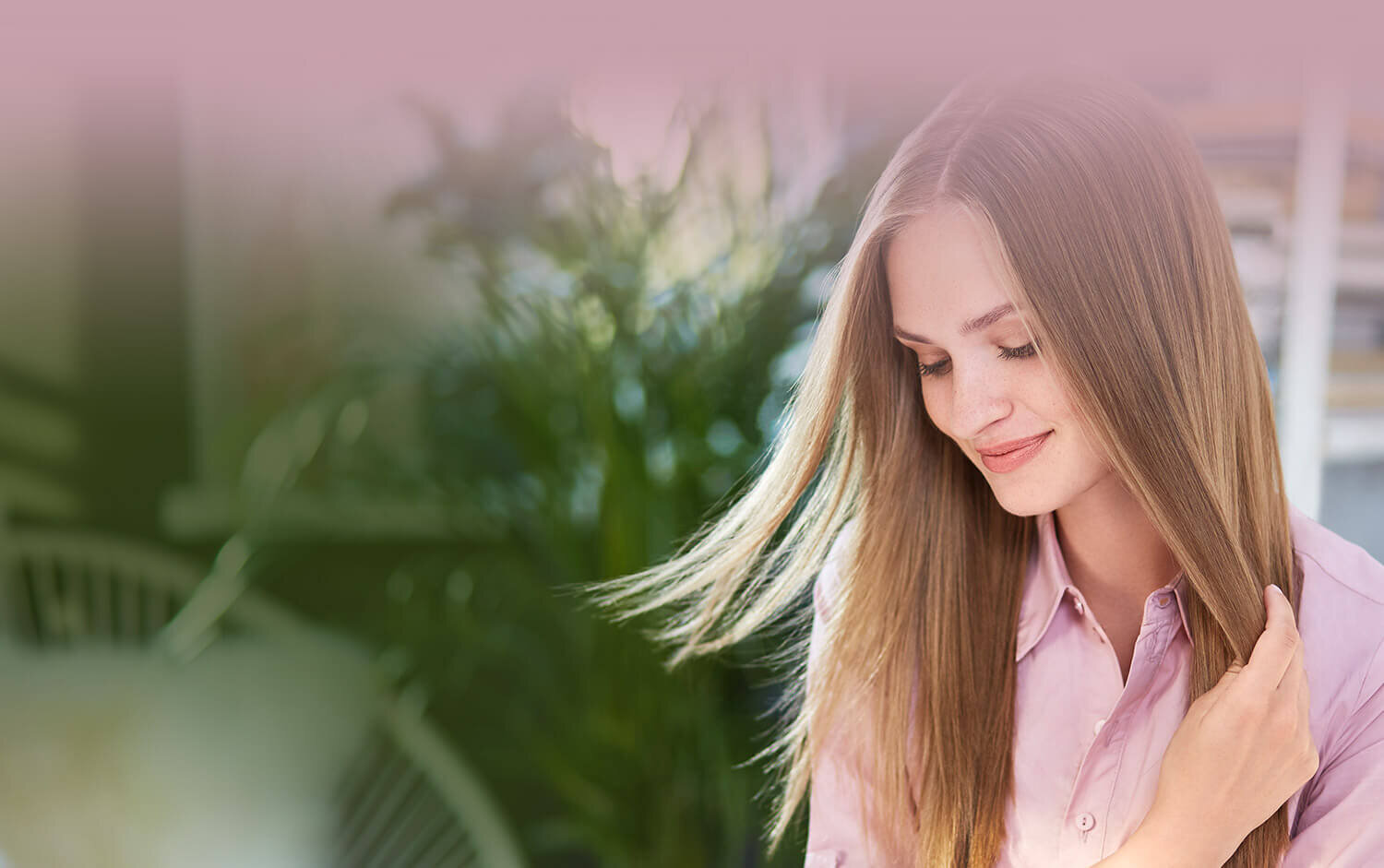 Osoba z długimi, prostymi włosami w różowej koszuli, w tle roślinność.