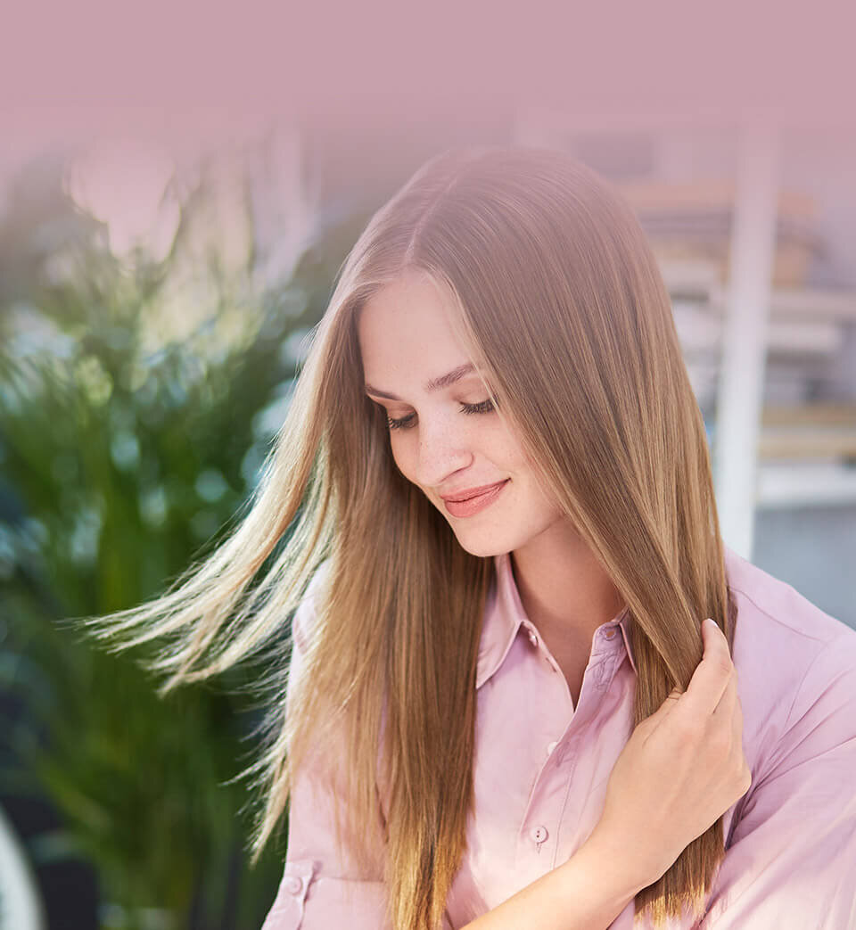 Osoba z długimi, prostymi włosami w różowej koszuli, trzymająca pasmo włosów. W tle widoczne są rozmyte rośliny i półki.