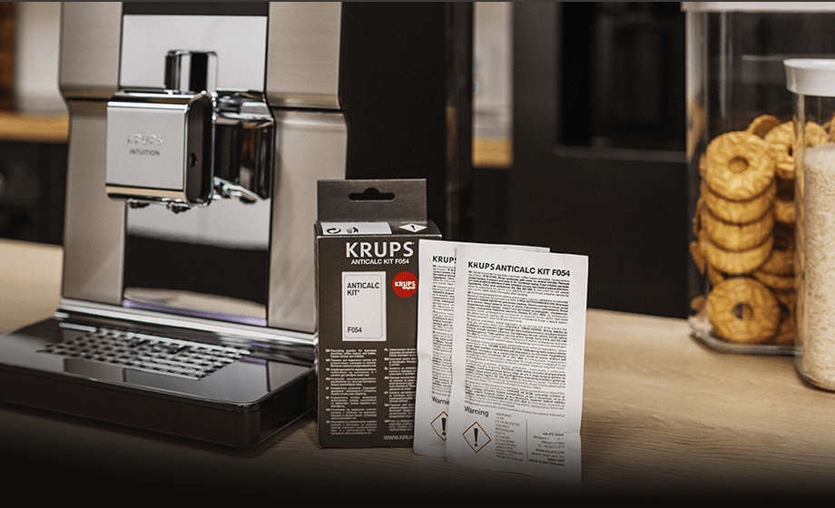 Na blacie znajduje się ekspres do kawy oraz opakowanie z napisem \'KRUPS ANTICALC KIT F054\'. Obok opakowania leżą dwie ulotki z tekstem \'KRUPS ANTICALC KIT F054\'.