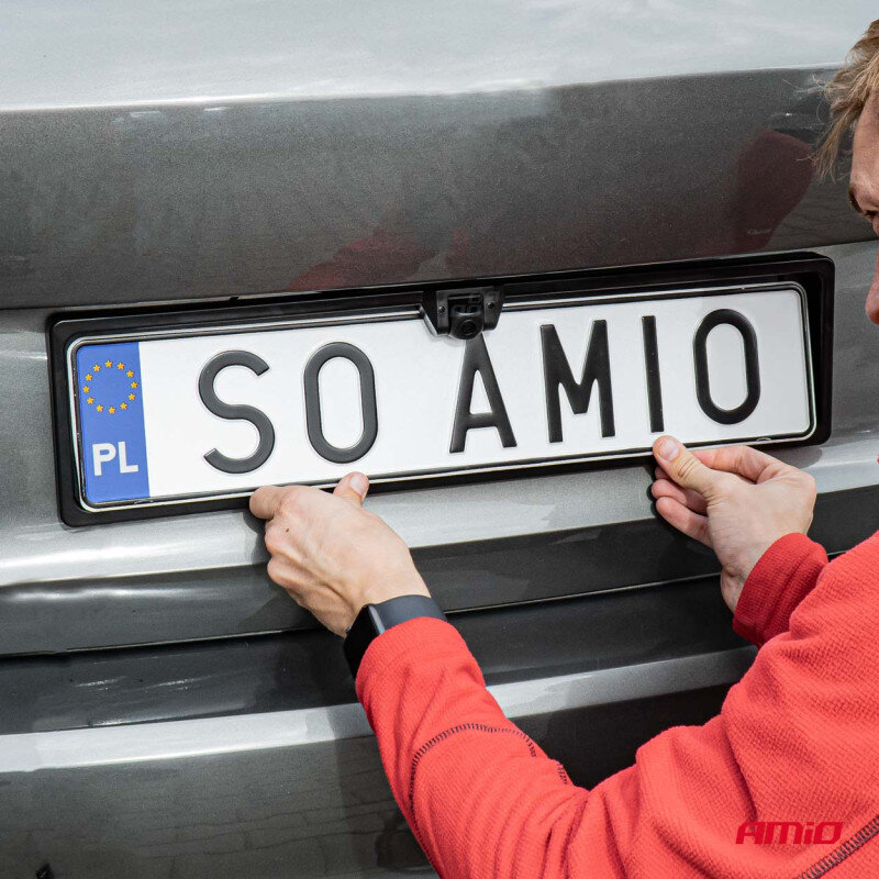 Osoba montuje tablicę rejestracyjną z napisem \'SO AMIO\' na samochodzie, w ramce z kamerą cofania. W prawym dolnym rogu widoczne jest logo \'AMiO\'.