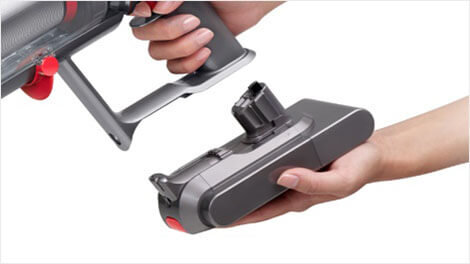 Osoba trzyma odkurzacz i wyjmuje z niego akumulator. Odkurzacz ma szary kolor z czerwonymi elementami.