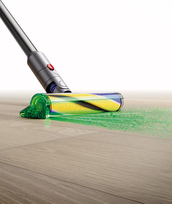 Na obrazie widoczna jest głowica odkurzacza z przezroczystą, zieloną osłoną, która zbiera zielony proszek z podłogi. Rura odkurzacza jest metaliczna z czerwonym przyciskiem.