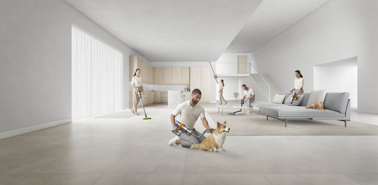 W przestronnym, nowoczesnym salonie kilka osób używa odkurzacza bezprzewodowego do sprzątania podłogi i kanapy. Na pierwszym planie mężczyzna odkurza sierść psa siedzącego na podłodze.