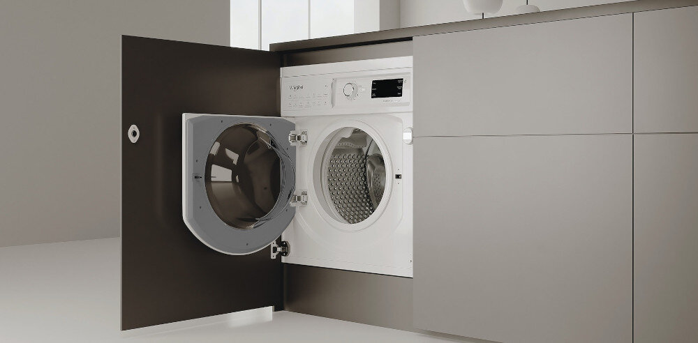 Pralka w zabudowie z otwartymi drzwiami, widoczny bęben i panel sterowania z pokrętłem oraz wyświetlaczem. Obok pralki znajdują się szafki kuchenne w ciemnym kolorze.