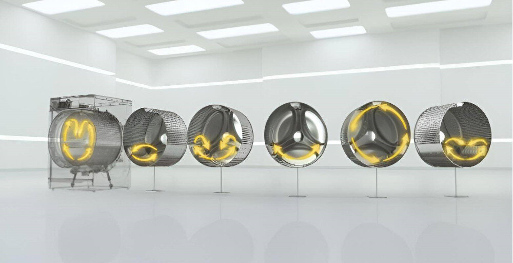 Obraz przedstawia sześć bębnów pralki ustawionych w rzędzie w jasnym pomieszczeniu. Na bębnach widoczne są żółte strzałki wskazujące kierunek obrotu.