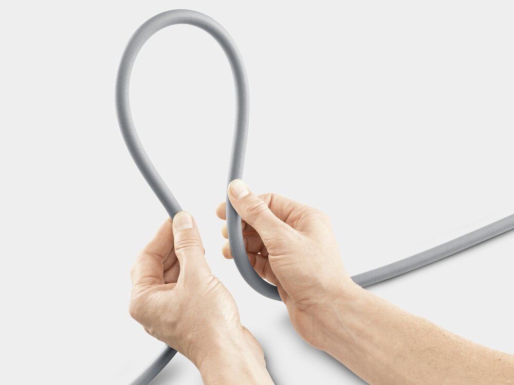 Dwie dłonie trzymają szary wąż ogrodowy, formując z niego pętlę. Tło jest jednolite i jasne.