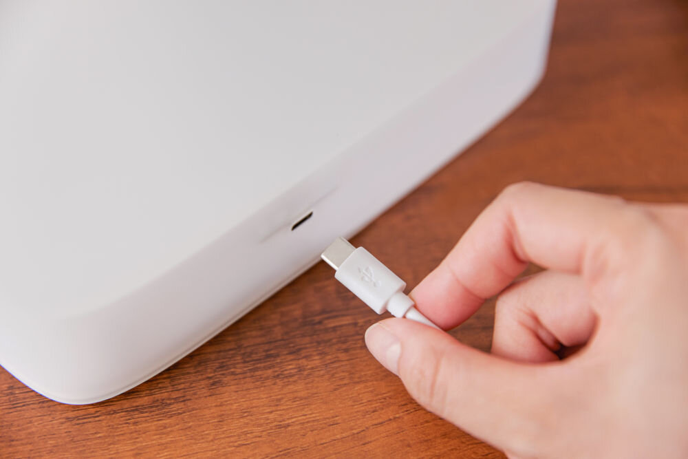 Osoba trzyma biały kabel USB-C, zbliżając go do portu w białym urządzeniu na drewnianym blacie.