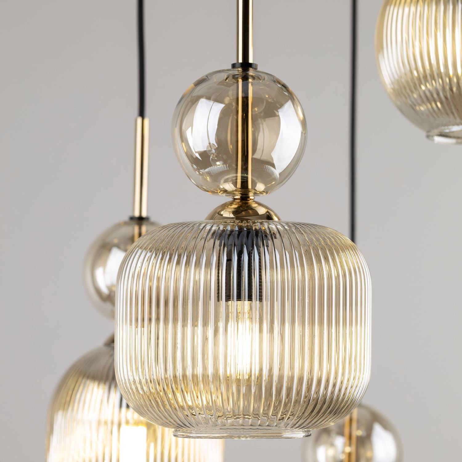 Zbliżenie na nowoczesną lampę wiszącą z przezroczystymi, żebrowanymi kloszami i złotymi elementami metalowymi. Lampa jest zawieszona na czarnych przewodach, a w tle widoczna jest neutralna, jasna przestrzeń.