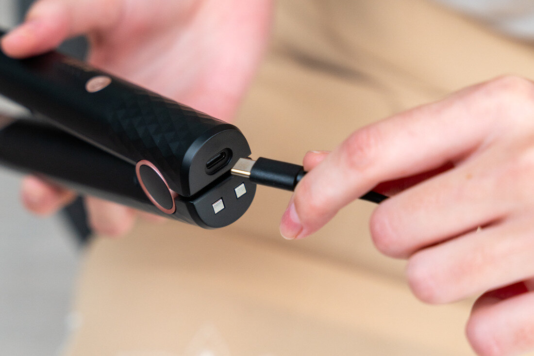 Osoba trzyma czarną prostownicę z teksturowaną powierzchnią, do której podłączany jest kabel USB-C. Na prostownicy widoczny jest okrągły przycisk.