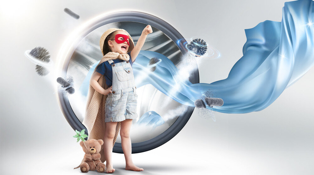 Dziecko w stroju superbohatera stoi przed dużym bębnem pralki, trzymając rękę w górze. Obok niego znajduje się pluszowy miś, a z pralki wydobywa się niebieska tkanina i efekty świetlne.