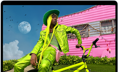 Osoba w jaskrawozielonym stroju siedzi na rowerze przed różowym budynkiem. W tle widoczny jest księżyc i ptaki na niebie.