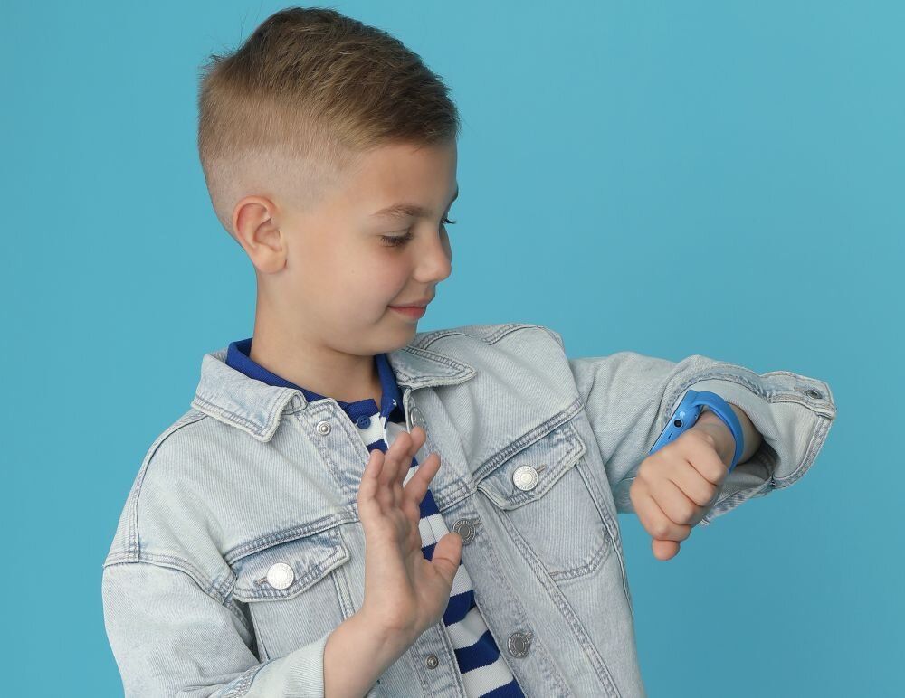Dziecko w jasnej kurtce patrzy na niebieski smartwatch na nadgarstku, na tle jednolitej, niebieskiej ściany.