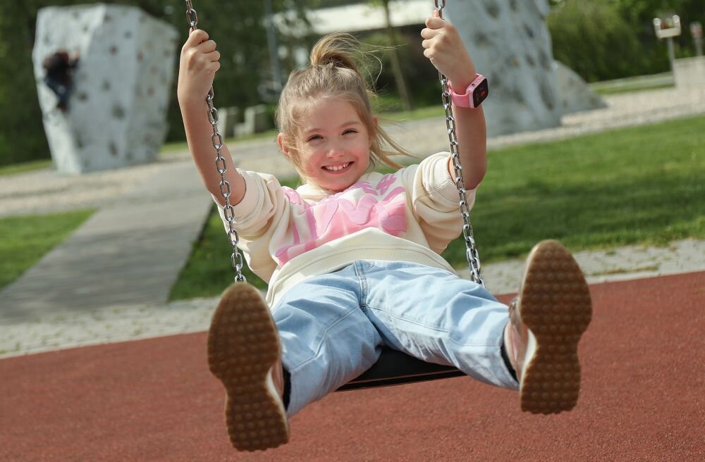 Dziecko siedzi na huśtawce na placu zabaw, ubrane w jasną bluzę i dżinsy, z różowym smartwatchem na nadgarstku. W tle widoczne są elementy placu zabaw, takie jak ścianka wspinaczkowa.