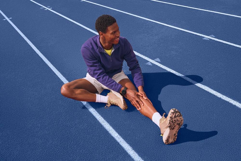 Osoba siedzi na niebieskim torze biegowym, rozciągając nogi. Na nadgarstku ma założony czarny smartwatch.