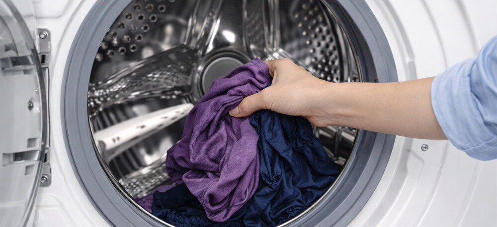 Osoba wkłada kolorowe ubrania do bębna pralki. Wnętrze pralki jest metaliczne i błyszczące.
