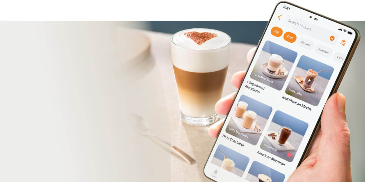 Osoba trzyma smartfon z aplikacją do wyboru przepisów na kawę, a w tle stoi szklanka z kawą z pianką i wzorem z kakao na wierzchu.