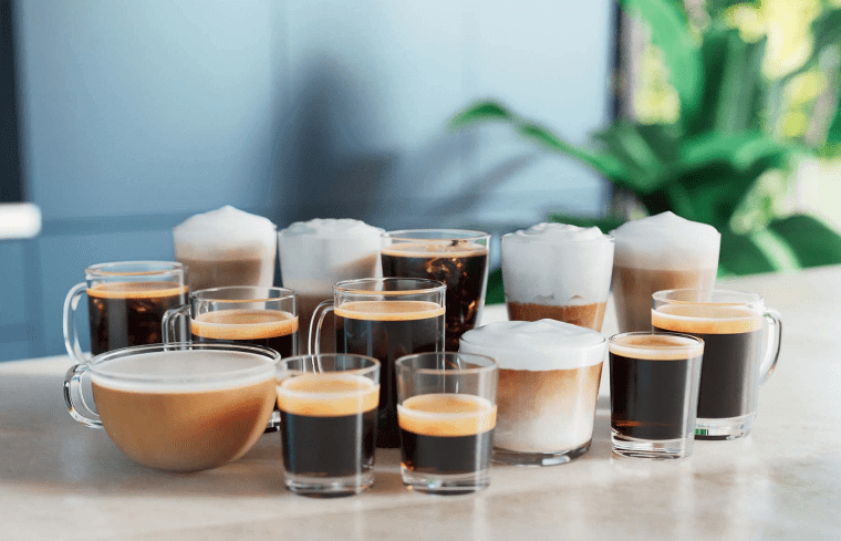 Na blacie znajduje się zestaw filiżanek i szklanek z różnymi rodzajami kawy, w tym kawy z pianką mleczną. W tle widoczne są rozmyte elementy wnętrza i roślinność.