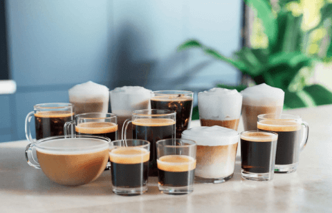 Na stole znajduje się kilka filiżanek i szklanek z różnymi rodzajami kawy, w tym kawy z pianką mleczną. W tle widoczne są rozmyte elementy zieleni.