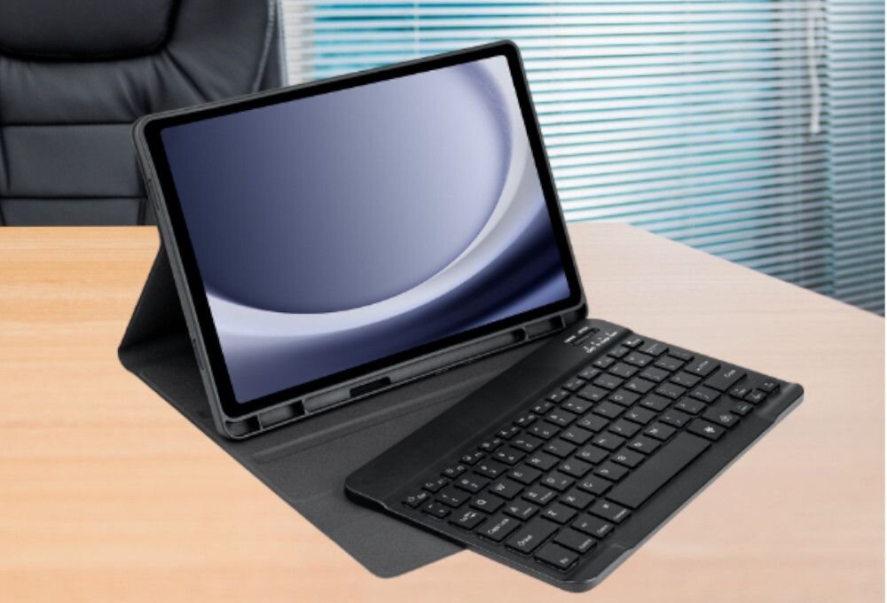 Na biurku znajduje się tablet w czarnym etui z klawiaturą. W tle widoczne są żaluzje i fragment fotela.