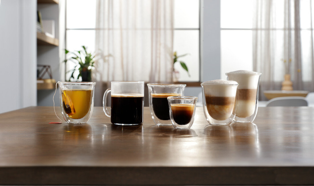 Na drewnianym stole stoi sześć szklanek z różnymi napojami, w tym herbata, kawa czarna, espresso i kawy z mlekiem. W tle widać rozmyte wnętrze z roślinami i zasłonami.