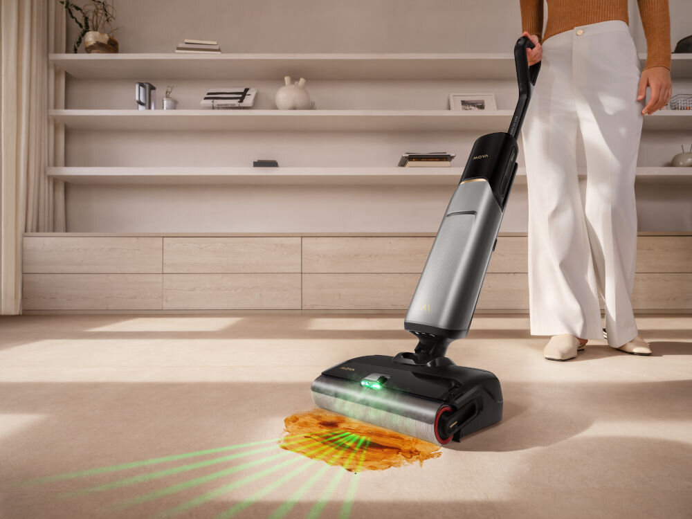 Osoba używa odkurzacza pionowego na jasnym dywanie, na którym znajduje się plama. Odkurzacz emituje zielone światło na powierzchnię dywanu.