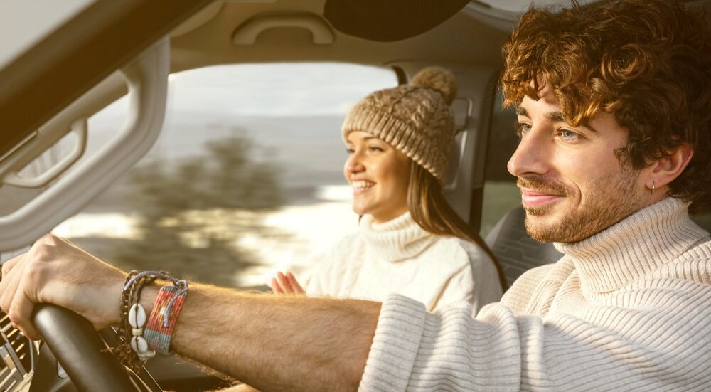Dwie osoby siedzą w samochodzie, ubrane w jasne swetry. Osoba po lewej stronie prowadzi pojazd, a osoba po prawej uśmiecha się.