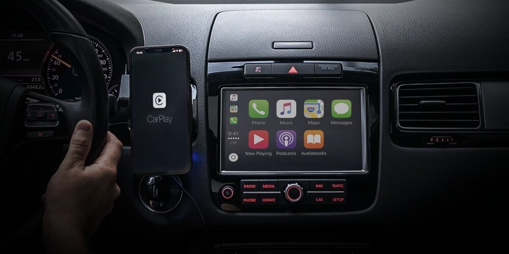 Wnętrze samochodu z widocznym ekranem systemu multimedialnego, na którym wyświetlane są ikony aplikacji takich jak Phone, Music, Maps, Messages. Obok znajduje się smartfon z logo CarPlay na ekranie, zamocowany w uchwycie.
