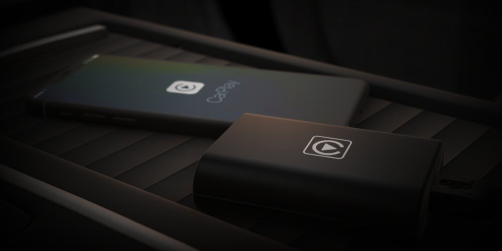 Na obrazie widoczny jest bezprzewodowy adapter samochodowy z logo oraz smartfon z wyświetlonym napisem \'CarPlay\'. Oba urządzenia leżą na ciemnej powierzchni.