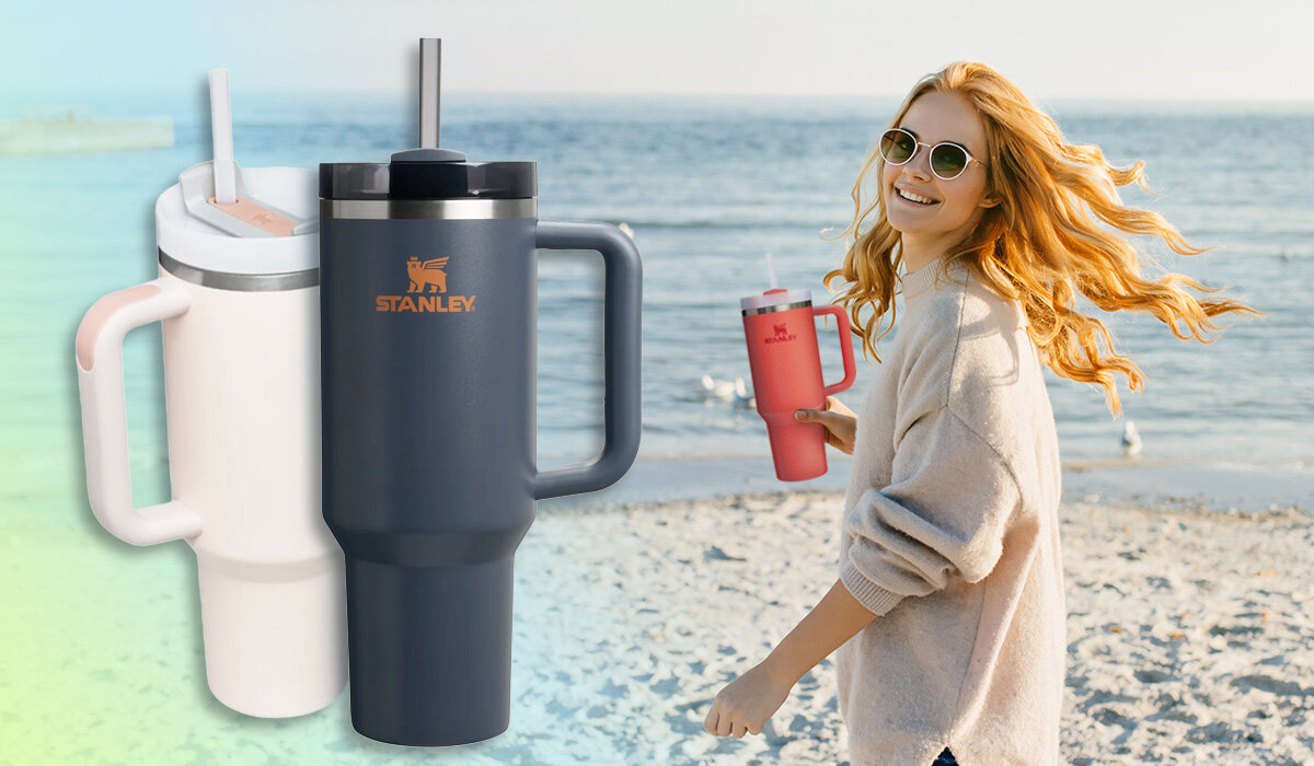 Na obrazie widoczne są trzy kubki termiczne marki Stanley w różnych kolorach oraz osoba trzymająca jeden z nich na plaży. Logo \'Stanley\' jest widoczne na kubkach.