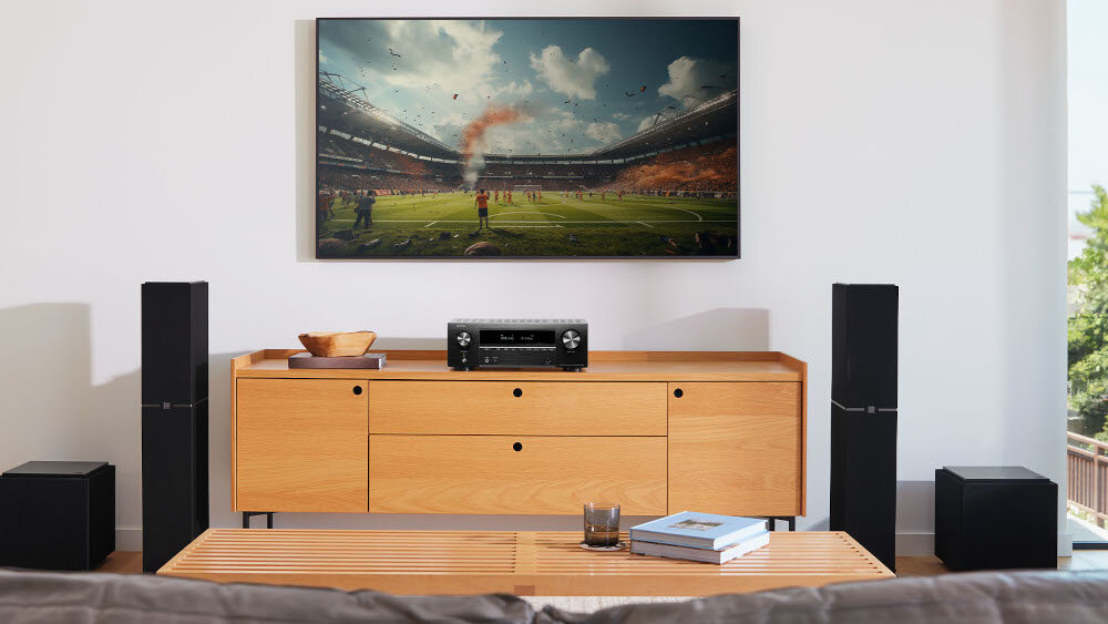 W salonie na drewnianej szafce stoi amplituner, a nad nim wisi telewizor wyświetlający mecz piłkarski na stadionie. Obok szafki znajdują się dwa głośniki, a na stoliku leży książka i szklanka.