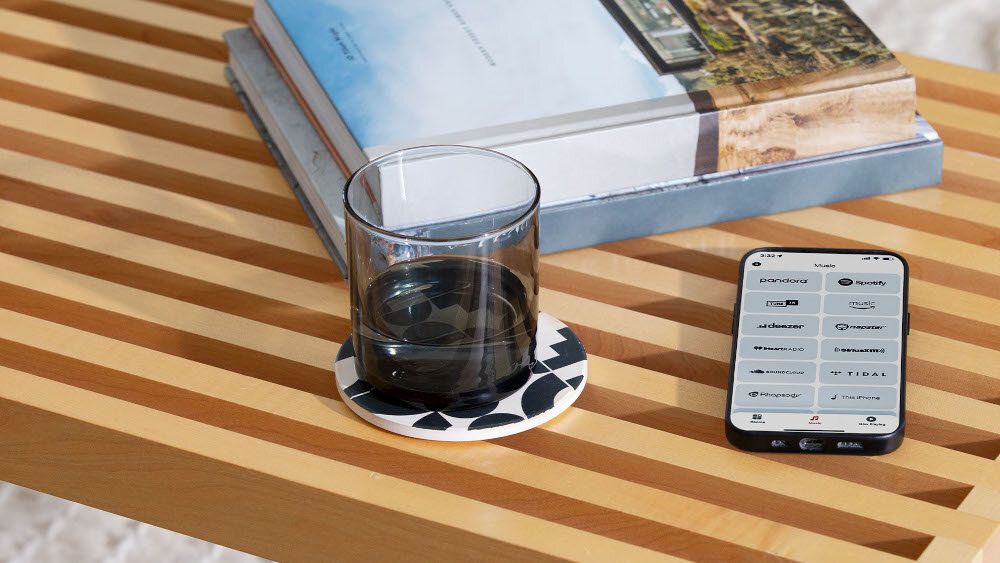 Na drewnianym stole znajduje się szklanka z ciemnym napojem na podstawce oraz smartfon z wyświetlonymi ikonami aplikacji muzycznych. Obok leży stos książek.