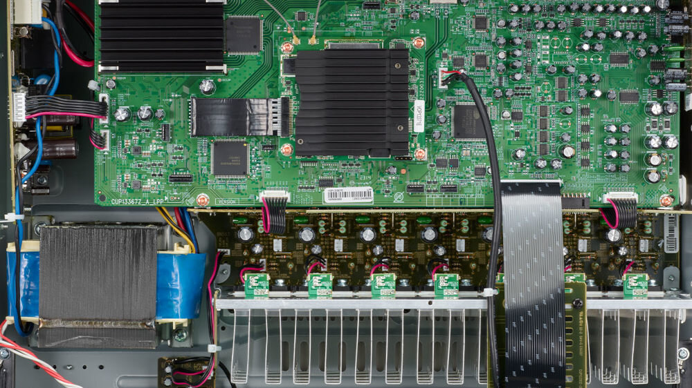 Wnętrze urządzenia elektronicznego z widoczną zieloną płytą główną, na której znajdują się różne komponenty elektroniczne i przewody. Widać również duży radiator oraz kilka złącz i kondensatorów.