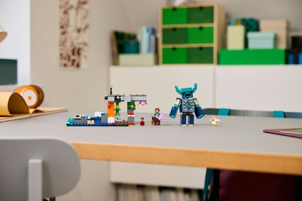 Na stole znajduje się zestaw klocków LEGO przedstawiający postacie i konstrukcje z gry Minecraft. W tle widoczne są półki z książkami i pudełkami.