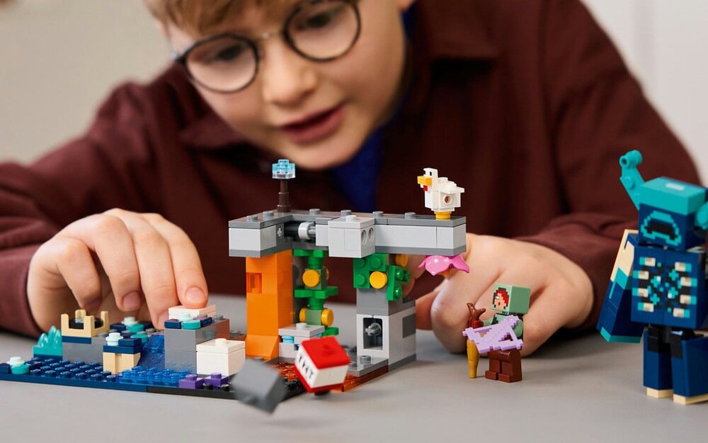 Dziecko bawi się zestawem klocków, który przedstawia scenę z gry Minecraft, z różnymi elementami konstrukcyjnymi i figurkami. Na stole widoczne są kolorowe klocki i postacie, w tym mały kurczak i inne elementy charakterystyczne dla gry.