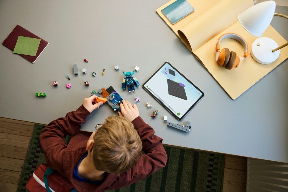 Na stole znajduje się zestaw klocków LEGO, z których część jest złożona, a obok leży tablet z wyświetlonym schematem budowy. W tle widoczne są słuchawki na stojaku oraz lampa.