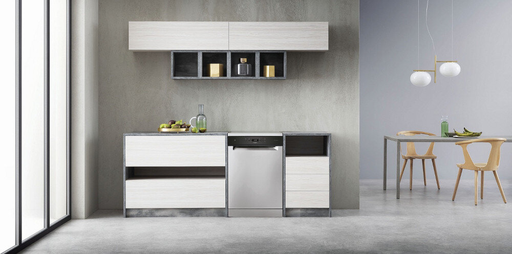 Nowoczesna kuchnia z zabudowaną zmywarką w kolorze inox, otoczona jasnymi szafkami. W tle widoczny jest stół z dwoma krzesłami oraz wiszące lampy.